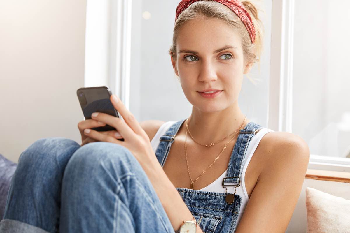 Pensive adorable woman in denim overalls, feels herself comfortable, uses modern smart phone for chatting at social networks, connected to wireless internet, being deep in thoughts. People, lifestyle